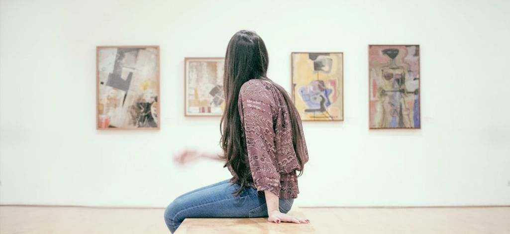 A woman sitting on a wooden bench in an art gallery, viewing abstract paintings displayed on the white wall in front of her.