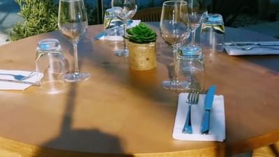 Table ronde extérieure avec vaisselle, verres à vin et plante en pot, entourée de chaises en osier.