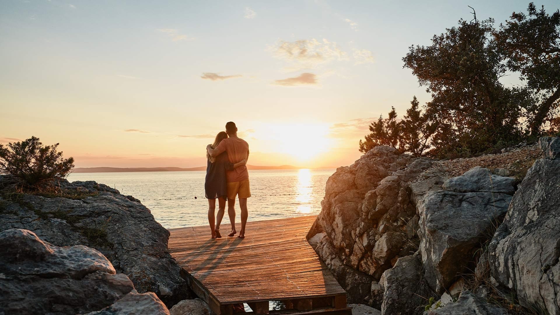 Par stoji na drvenom mostu iznad kamenja i gleda zalazak sunca iznad mora.