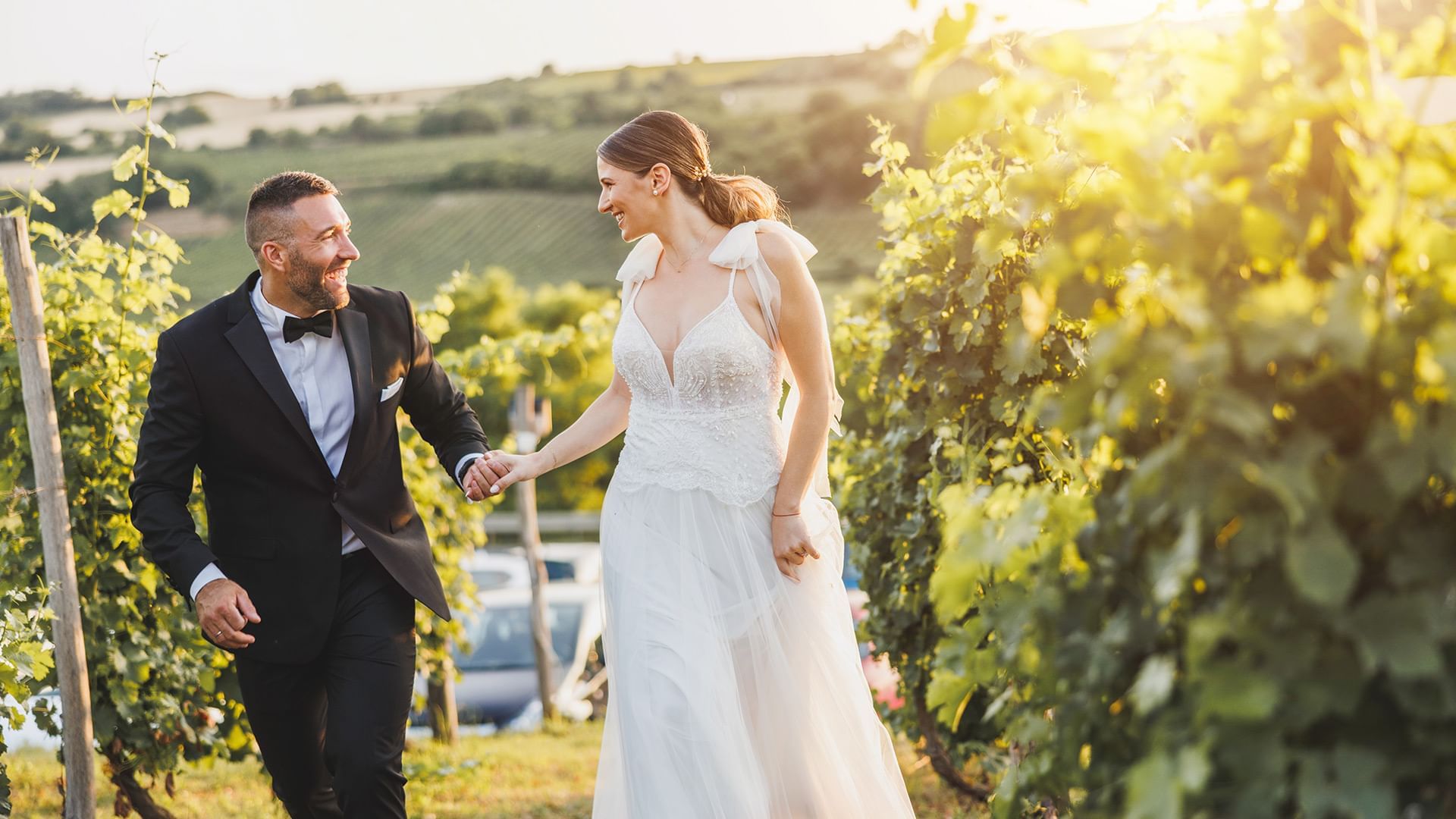 Wedded couple sharing a joyful moment, walking hand-in-hand through a sunlit vineyard near Coast Penticton Hotel