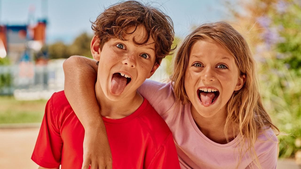 Zwei Kinder, ein Junge und ein Mädchen, stehen lachend und mit herausgestreckten Zungen nebeneinander.