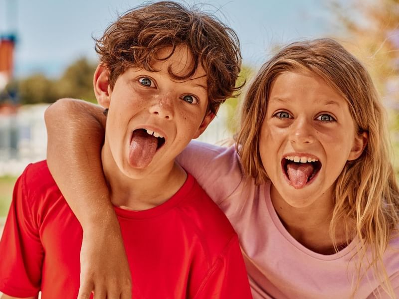 Due bambini, un ragazzo e una ragazza, sorridono con la lingua fuori e le braccia incrociate.