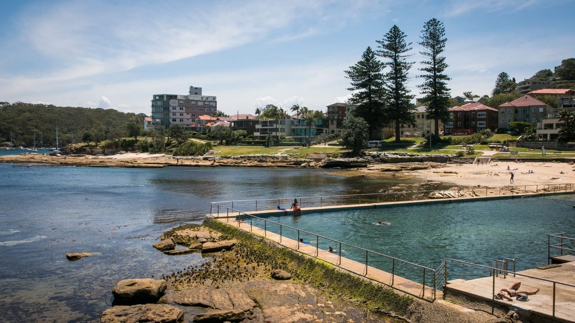 Fairlight Rockpool near Elysium Manly shows people swimming in a pool by a concrete path under a clear blue sky
