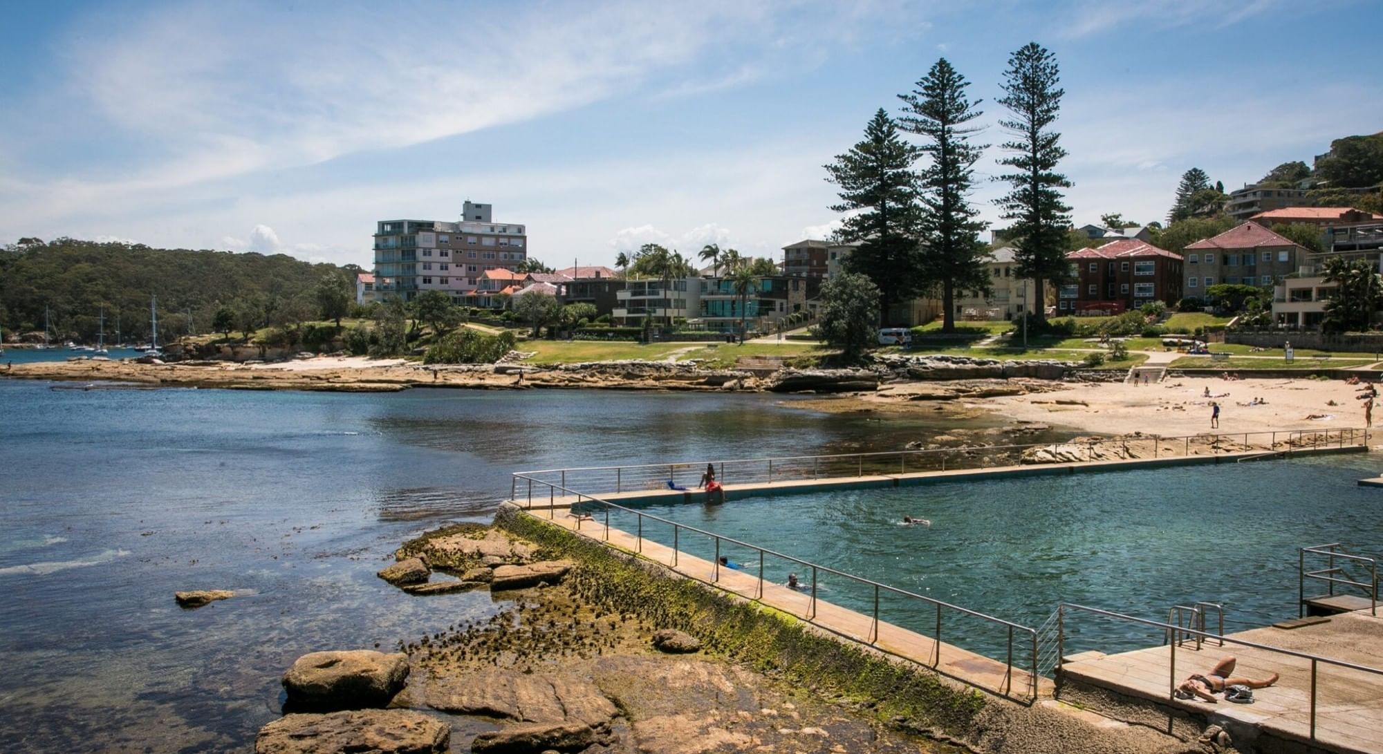 Fairlight Rockpool at Elysium Hotels is beside a rocky shore and ocean with swimmers and buildings behind a park