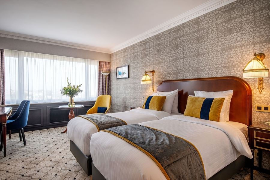 Twin beds with wood headboards and yellow armchairs by a bright window at Hotel Barsey by Warwick