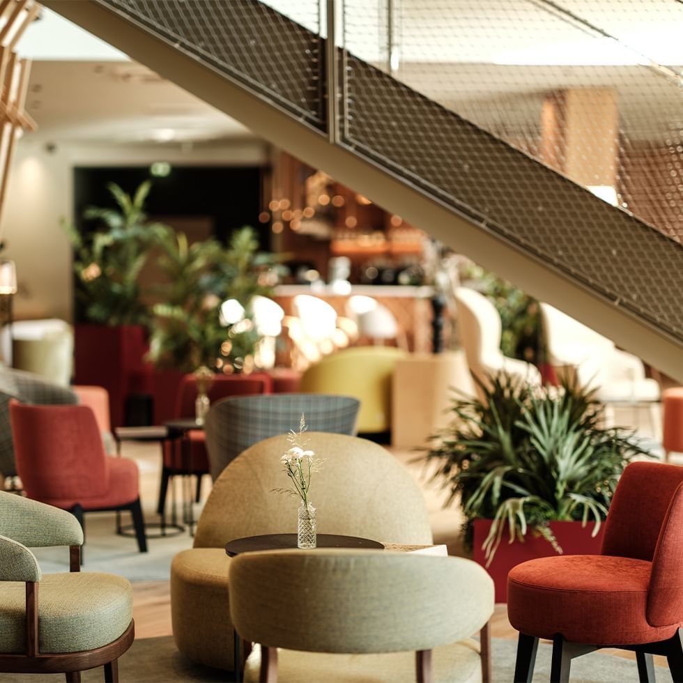 Sezione di un bar con diverse sedie colorate, piante, e un tavolino con un vaso di fiori.