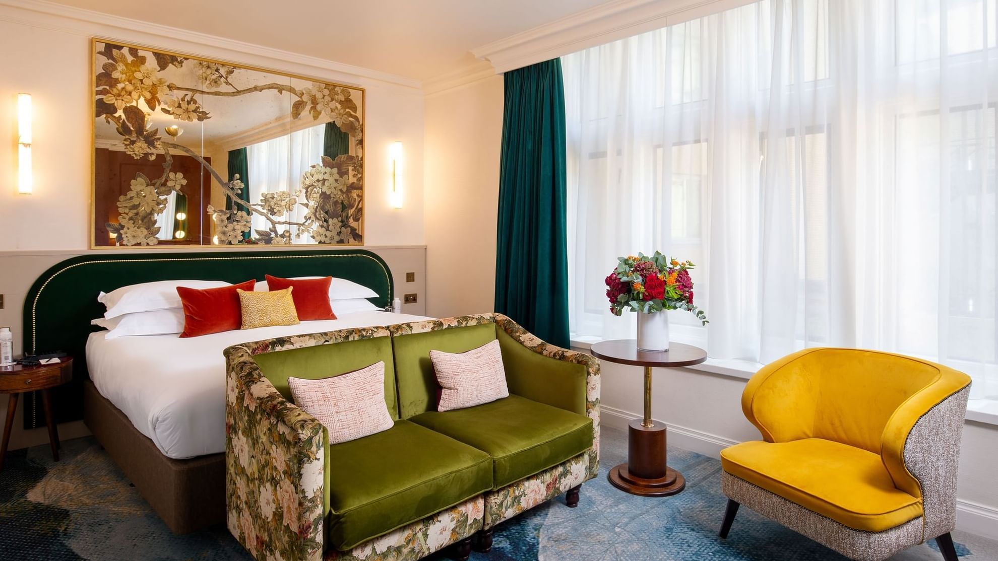 Elegant bedroom with large bed, green couch, yellow chair, floral decor, and large window with white curtains.
