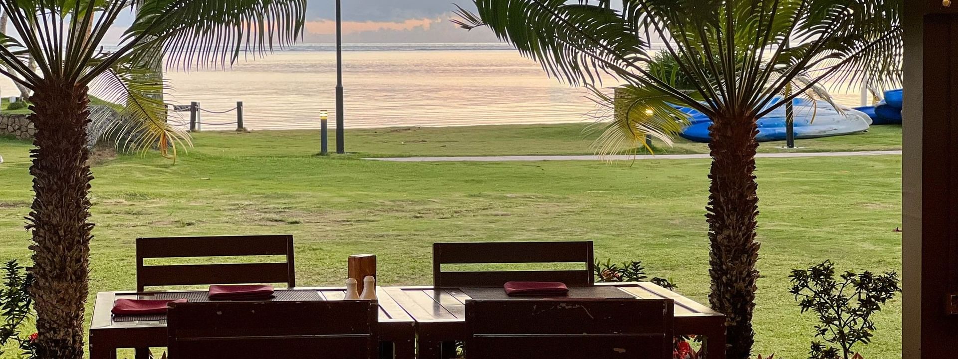 Wooden dining set under palm trees by a green lawn with ocean view at The Naviti Resort in Korolevu