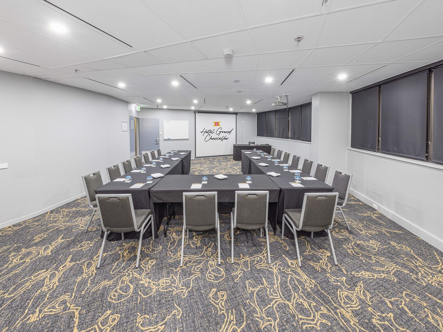 A conference room arranged in a U-shape layout with black tablecloths, grey chairs, and a projector screen displaying the hotel logo.