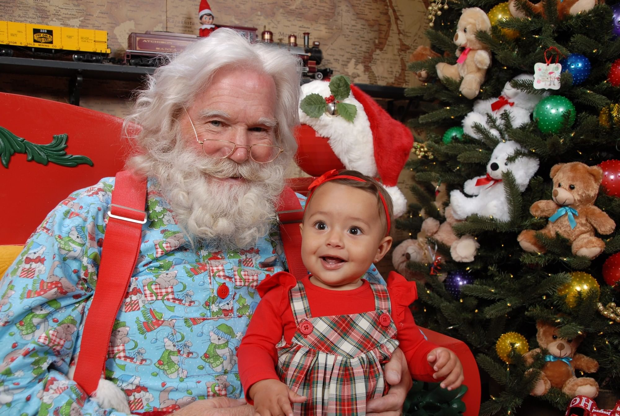 A casual Santa in suspenders holds a smiling baby girl on his lap beside a Christmas tree at ICON Park.