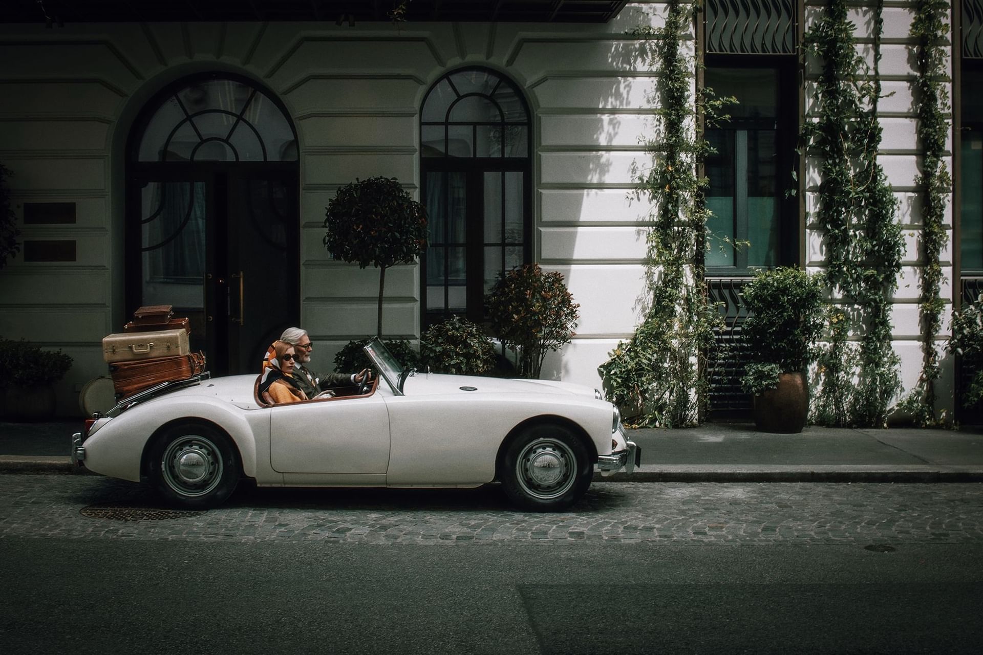 Couple driving a vintage car with leather luggage on a cobblestone street near Hotel Motto Vienna