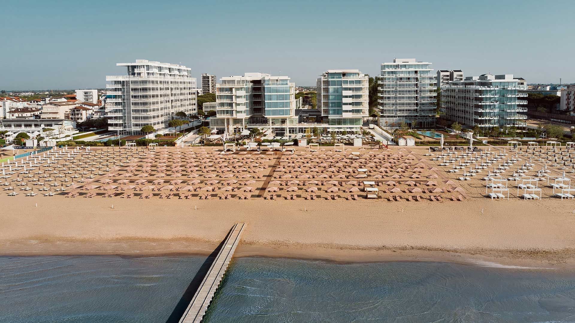Luftaufnahme eines Strandes mit vielen Sonnenschirmen und Liegestühlen, umgeben von Gebäuden.