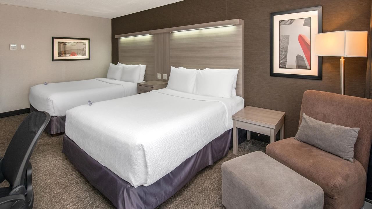 Modern hotel room with two queen beds, white linens, brown upholstered chair, and contemporary artwork at Coast Calgary University Area Hotel by APA