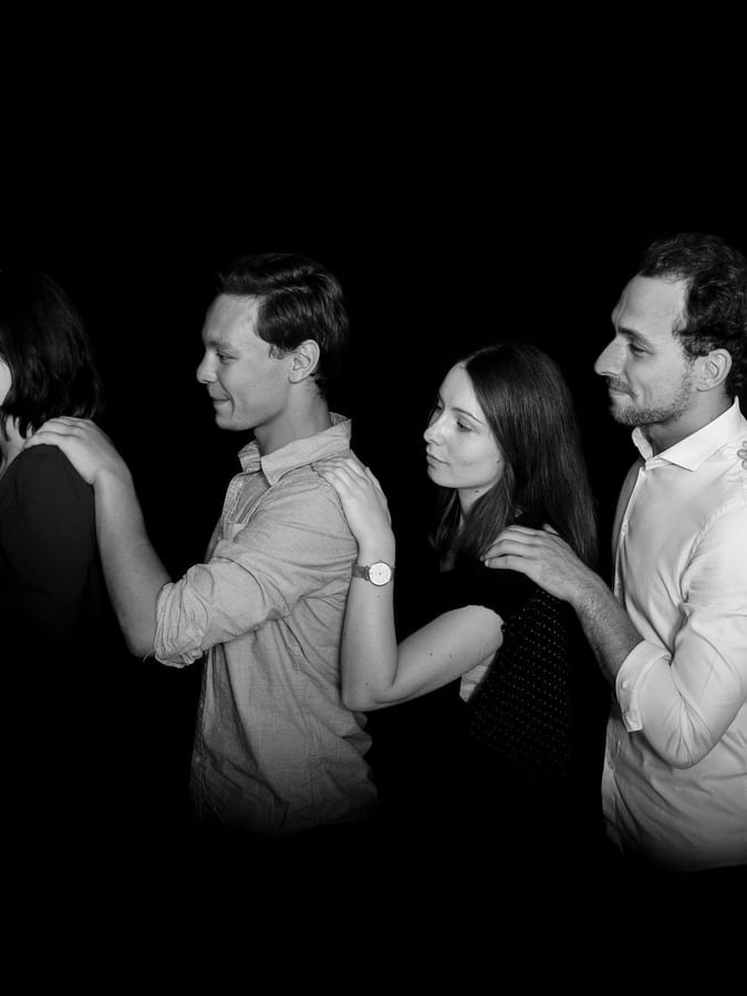 Five smiling people stand in a row against a black background, each resting a hand on the next person's shoulder