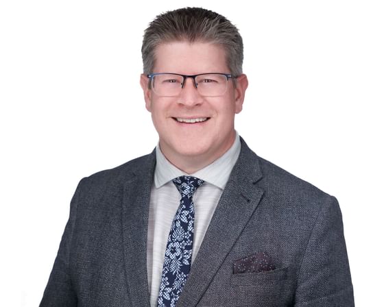 A man in a gray suit and blue tie, wearing glasses, smiles for the camera.