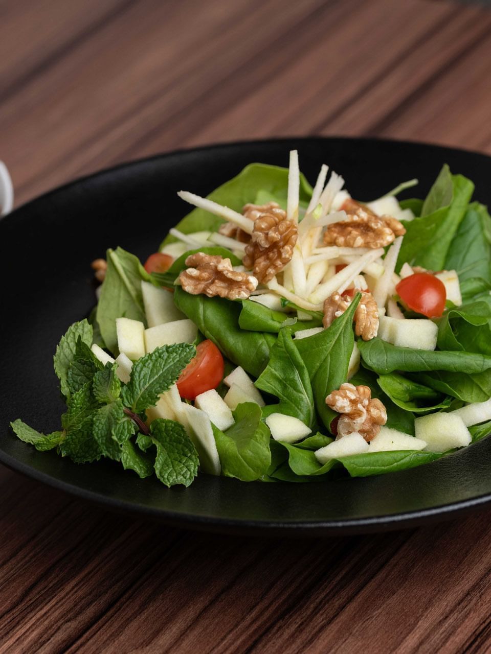Spinach salad with walnuts and apples in a black bowl by two small cups of dressing in KVNT at Warwick Al Jubail