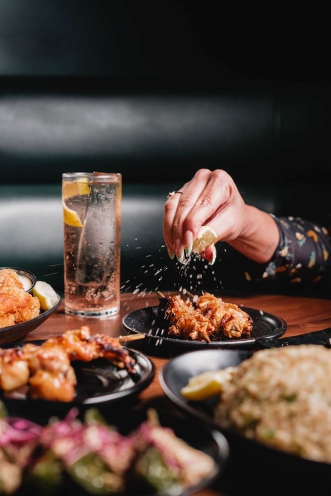 A table with multiple dishes on black plates with a cocktail and a hand squeezing a lemon wedge onto food. 