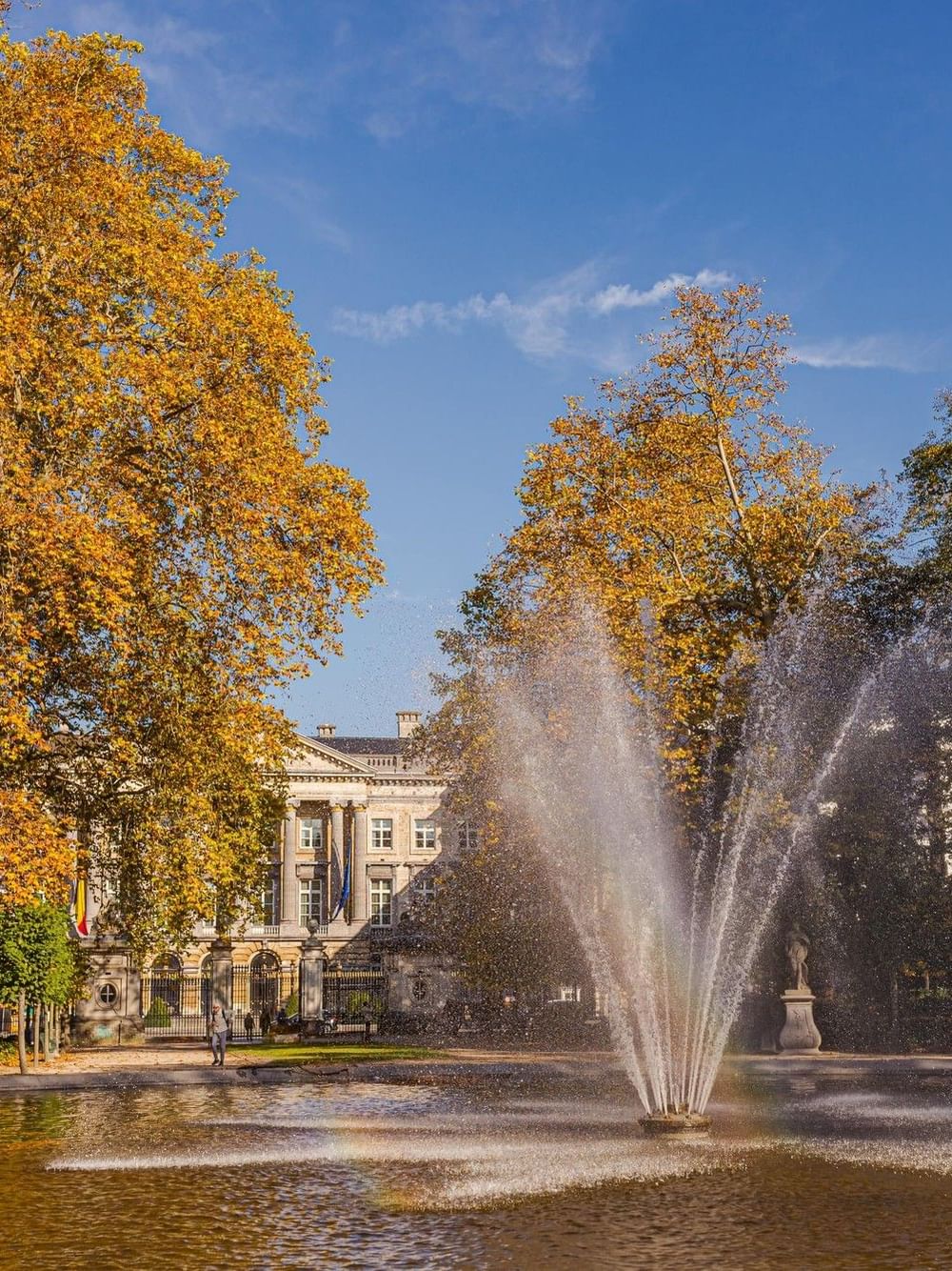 Fountain spraying water in a park surrounded by golden autumn trees and a historic building under a blue sky in Brussels.