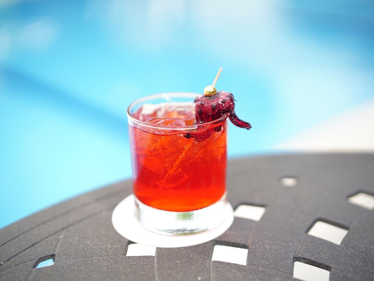 Red cocktail garnished with a dried hibiscus flower on a dark table by the pool at Pool Bar, Courtleigh Hotel & Suites