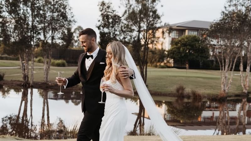 Bride and groom holding champagne glasses by a lake near Mercure Kooindah Waters