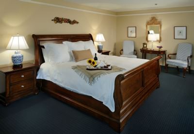 Spacious bedroom featuring a wooden sleigh bed with sunflowers and a wine tray beside armchairs at The Simsbury Inn