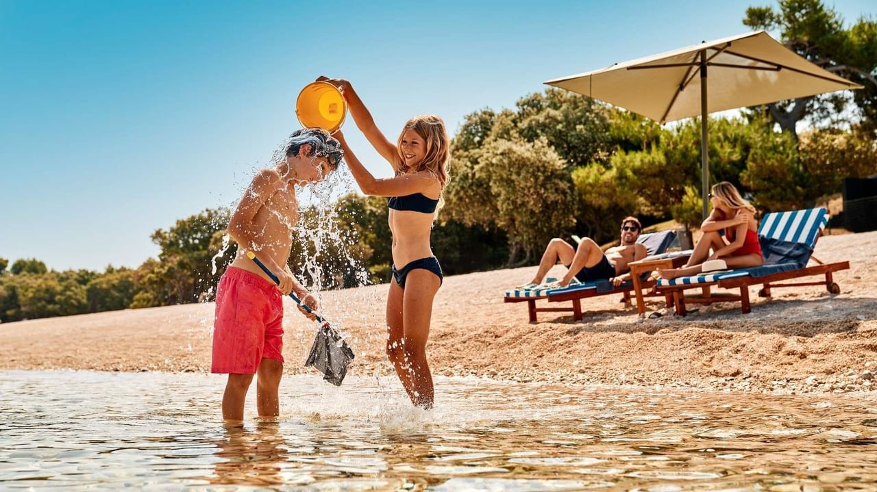 Bambini che giocano in acqua su una spiaggia, con ombrellone e lettini sullo sfondo.