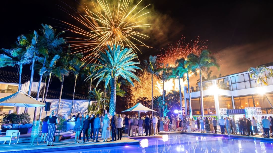Guests gather by a resort pool at night under fireworks, with illuminated palm trees and a building in the background.