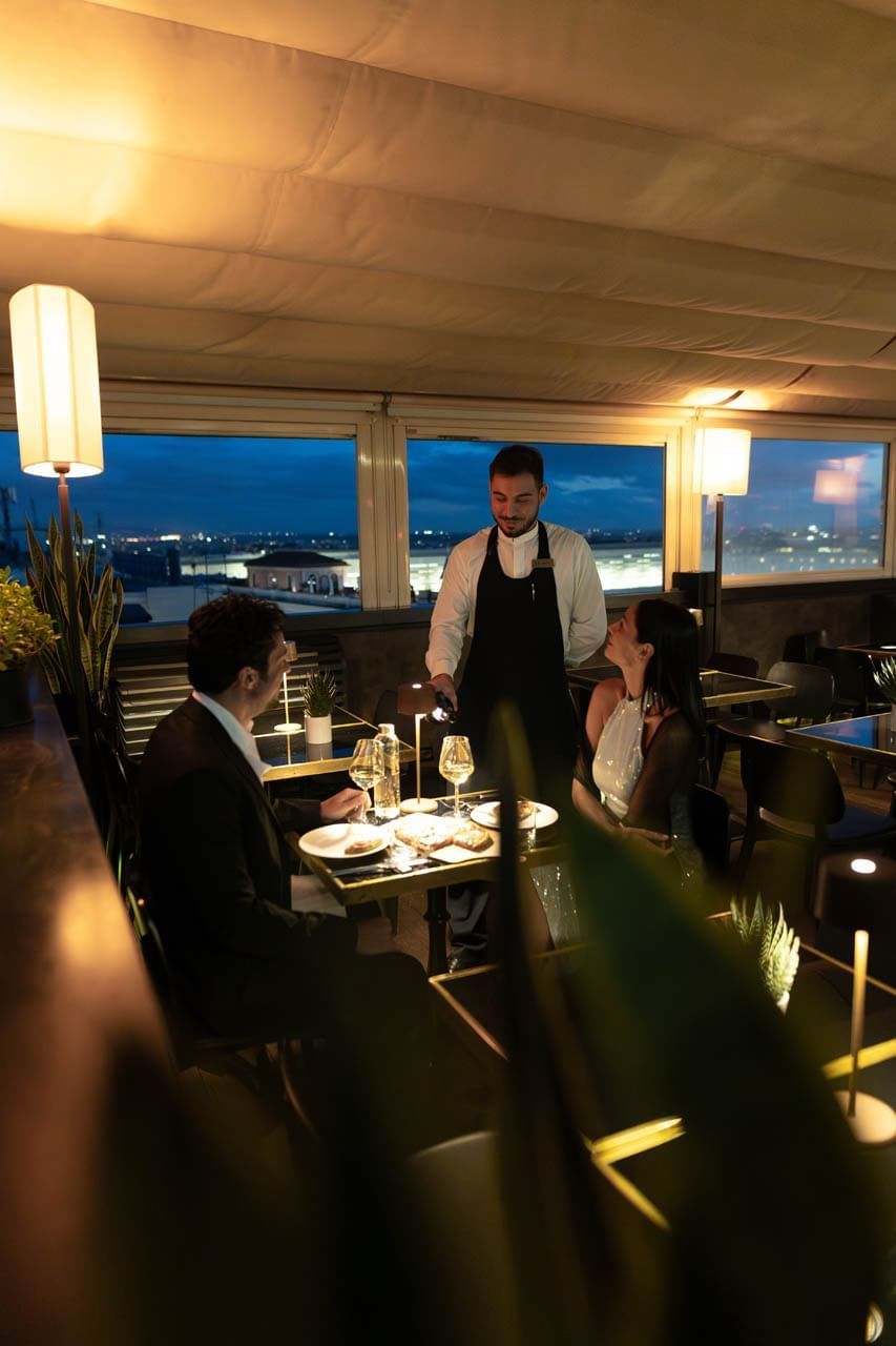 Waiter serves elegant dinner to couple at Terrazza Cielo, with scenic windows and soft dusk lighting at The Independent
