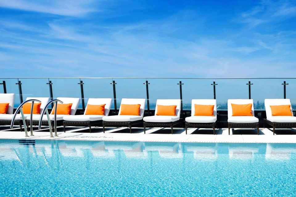 Rooftop swimming pool with row of white sun loungers and orange pillows under blue sky at Hotel X Toronto terrace
