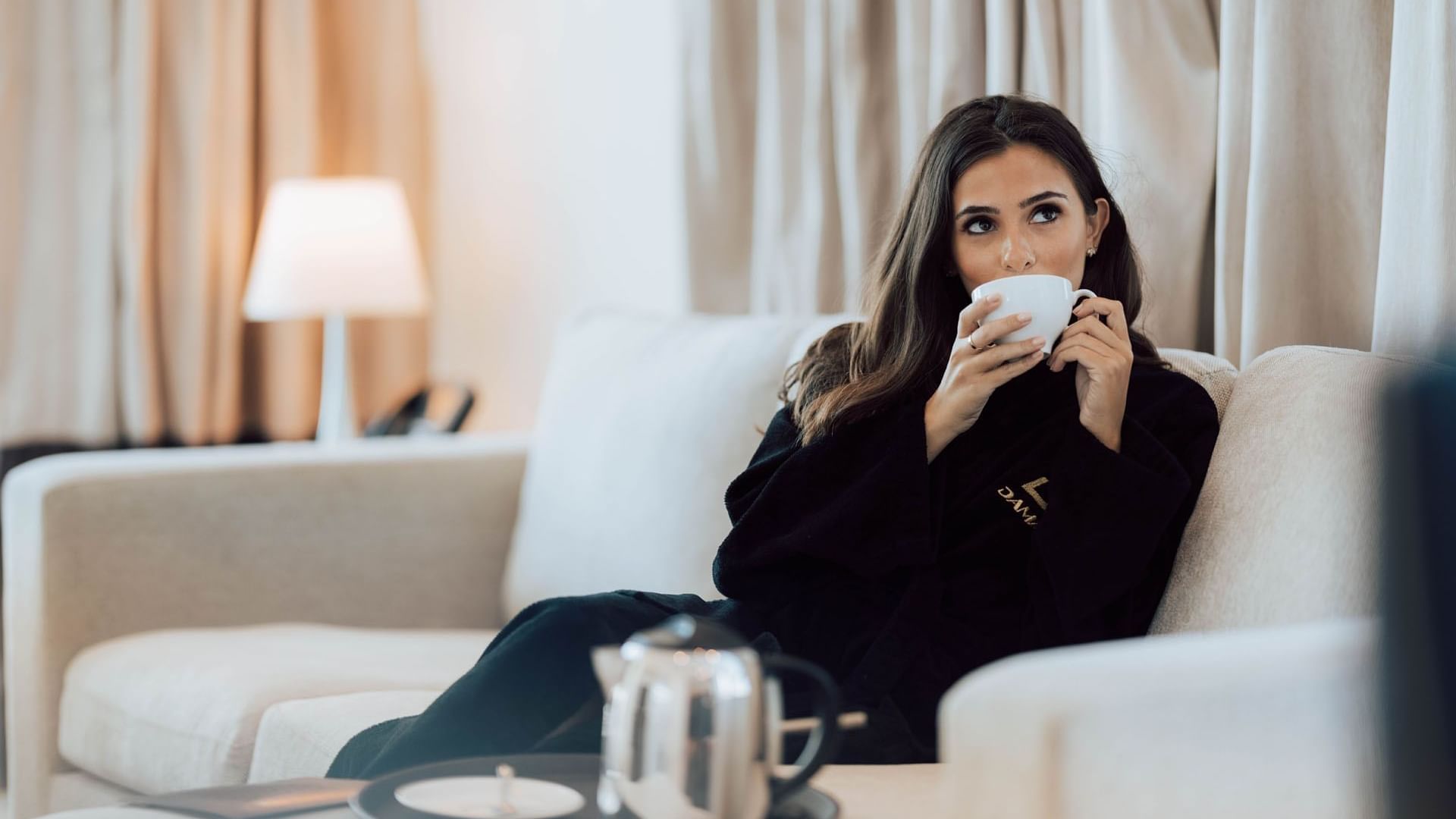 A lady drinking a cup of tea on a cozy couch in a hotel room at DAMAC Maison Distinction