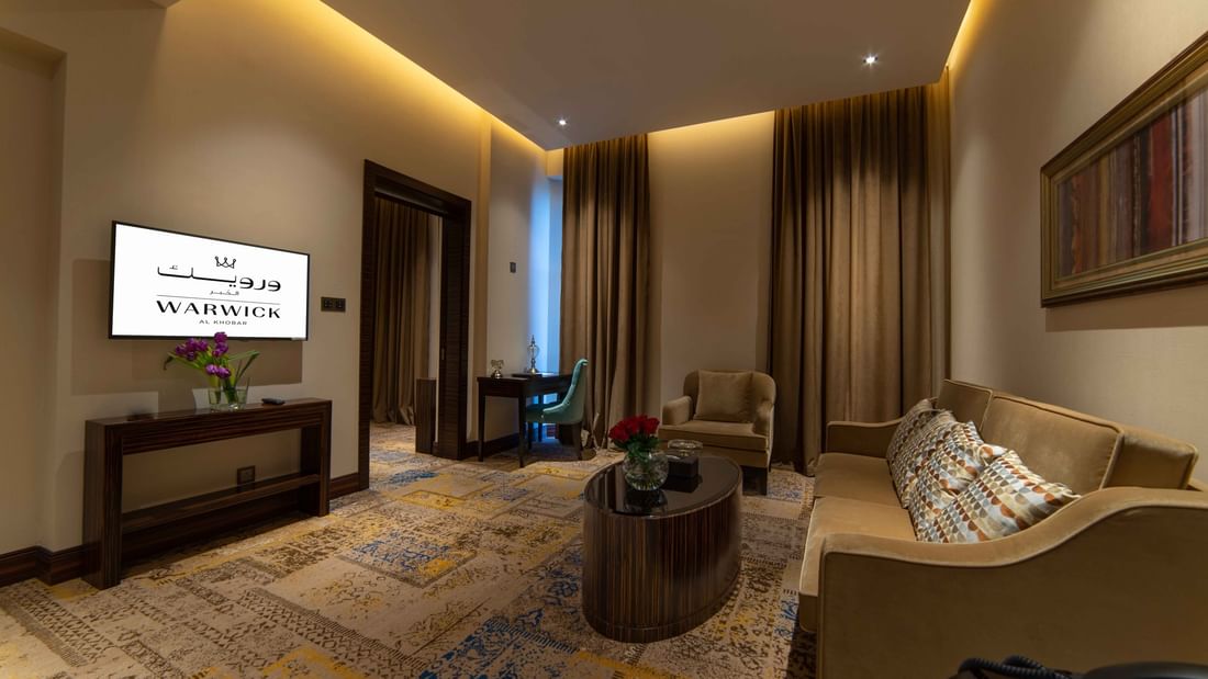 Gray sofas by an oval coffee table & turquoise desk chair on a vintage-patterned rug in Executive Room at Warwick Al Khobar