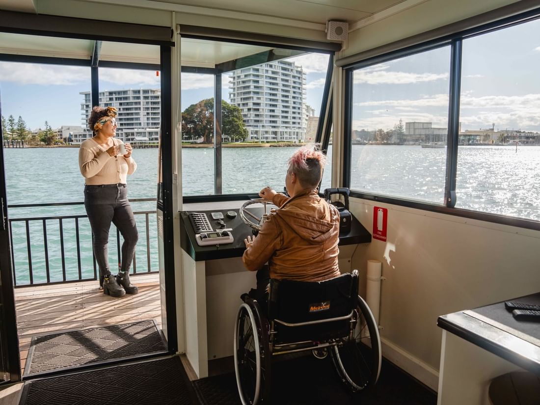 Guest in a wheelchair steers a boat, with another guest holding a mug on the deck, near The Sebel Mandurah