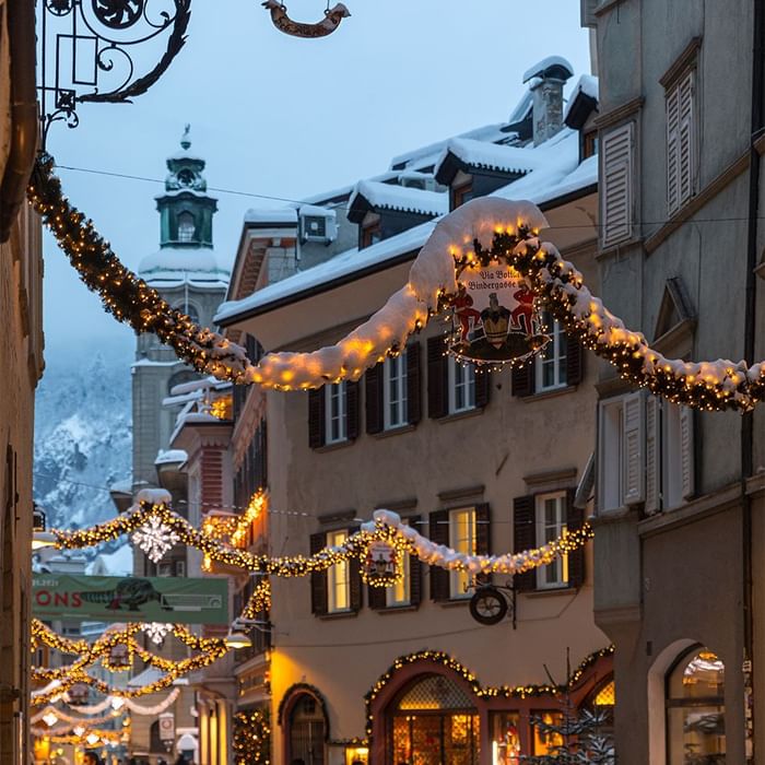 Strada decorata per Natale con luci e neve, edifici storici e tetti innevati sotto un cielo crepuscolare.