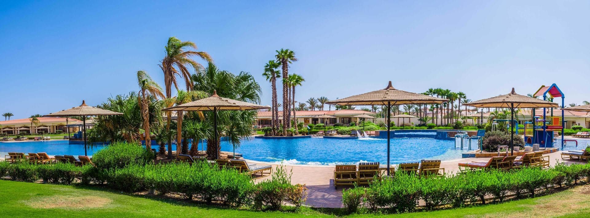 Pickalbatros Golf Beach Resort pool area with shady umbrellas, lounge chairs, palm trees & children's play area in distant
