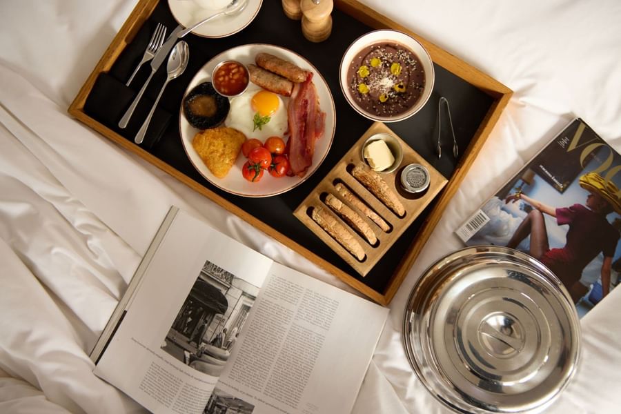 Tray with breakfast food, a magazine, and a silver lid on a bed at The May Fair Hotel London
