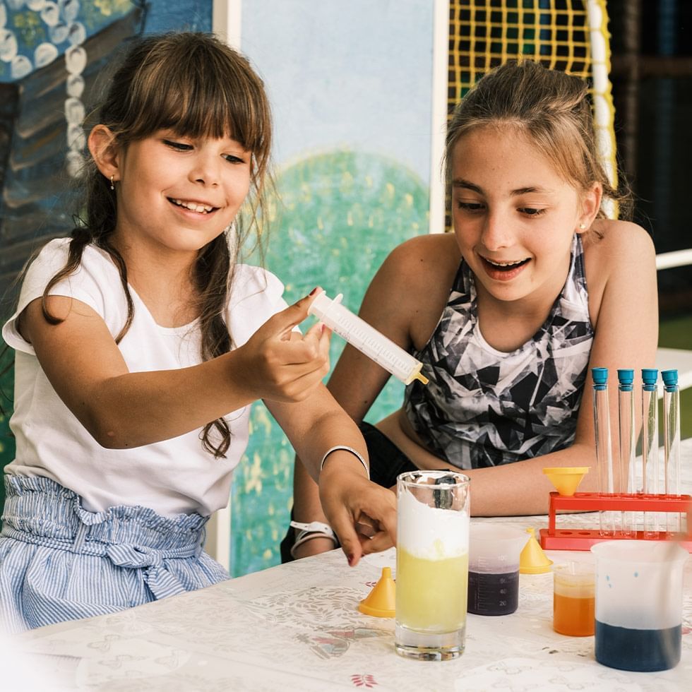 Due bambine sorridenti in un laboratorio, una sta aggiungendo una sostanza a un liquido in un bicchiere.
