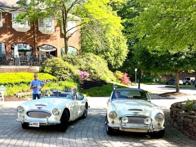 The Simsbury Inn driveway with two white vintage convertible sports cars and passengers posing in front of the entrance
