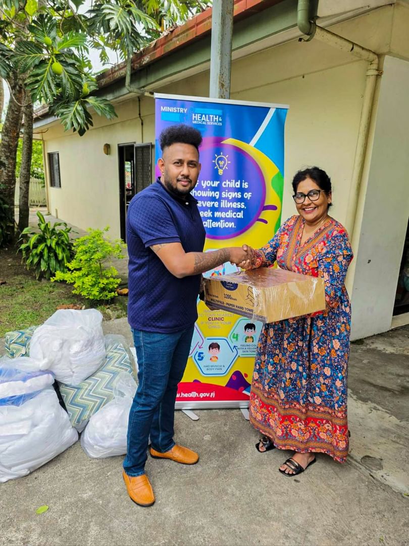 Two people shaking hands during a linen donation handover outside a health center with Ministry of Health signage visible.