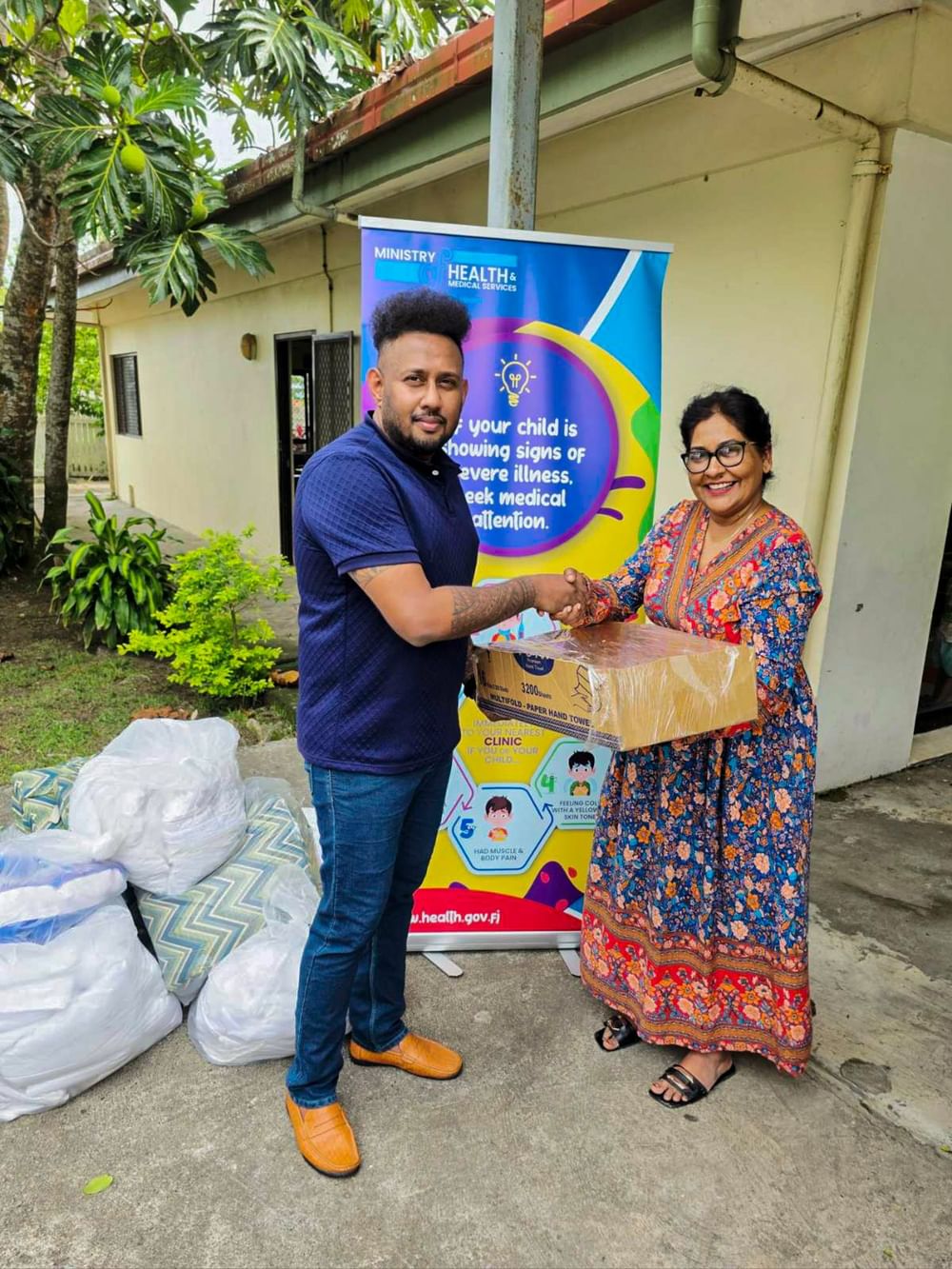 Two people shaking hands during a linen donation handover outside a health center with Ministry of Health signage visible.