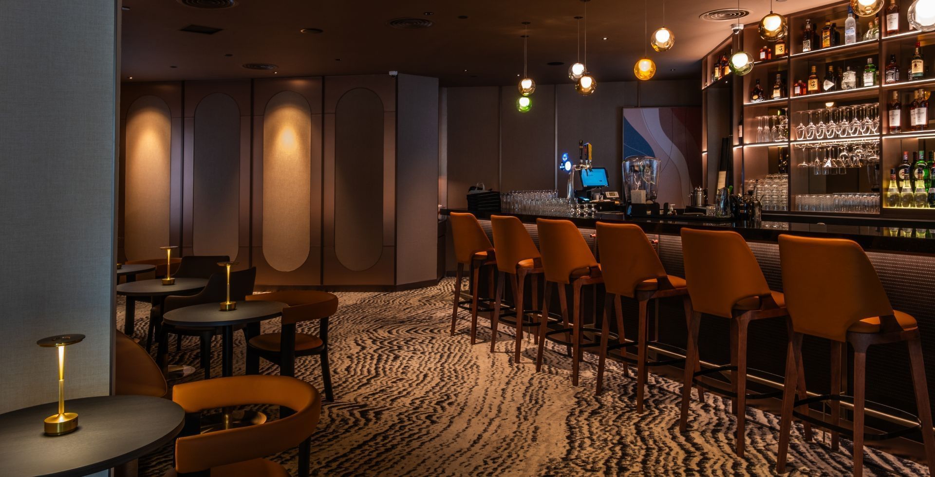 The dimly lit Tux Bar & Lounge with orange stools and patterned carpet offers a cozy setting at Carlton Hotel Singapore