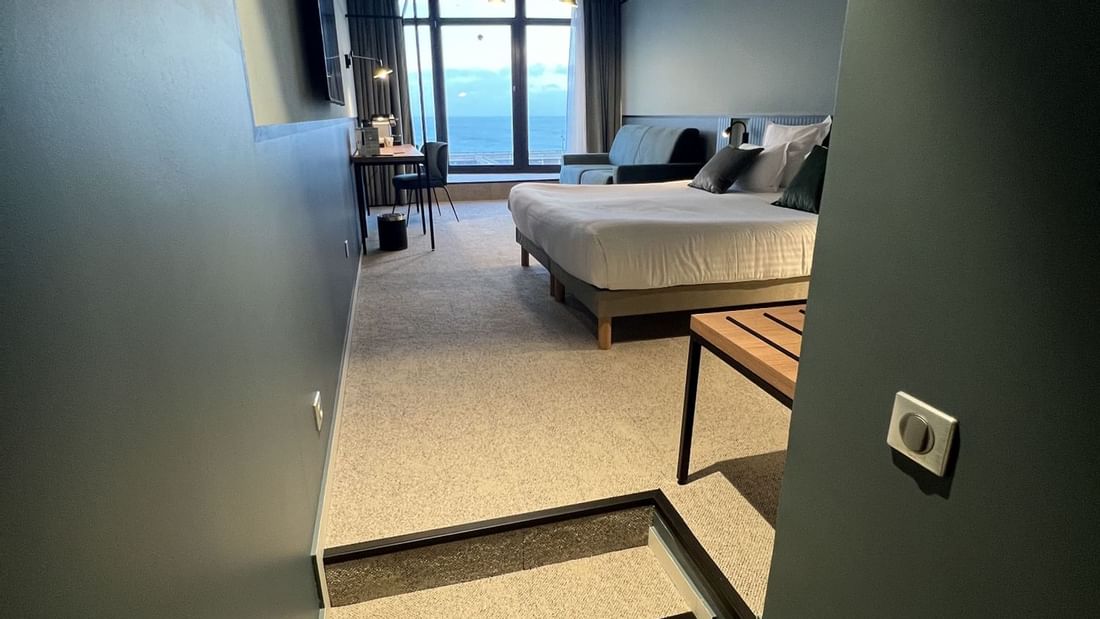 Vue en plan d'une chambre d'hôtel avec lit, bureau, canapé et vue sur l'océan.