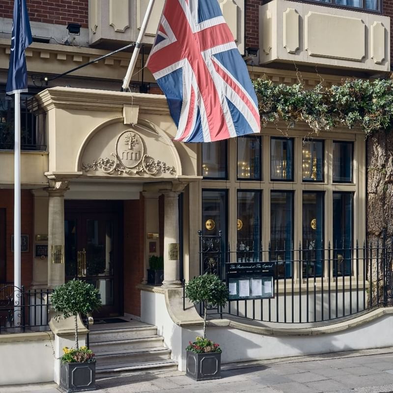 The entrance of a Capital Hotel London adorned with a British flag and surrounded by greenery