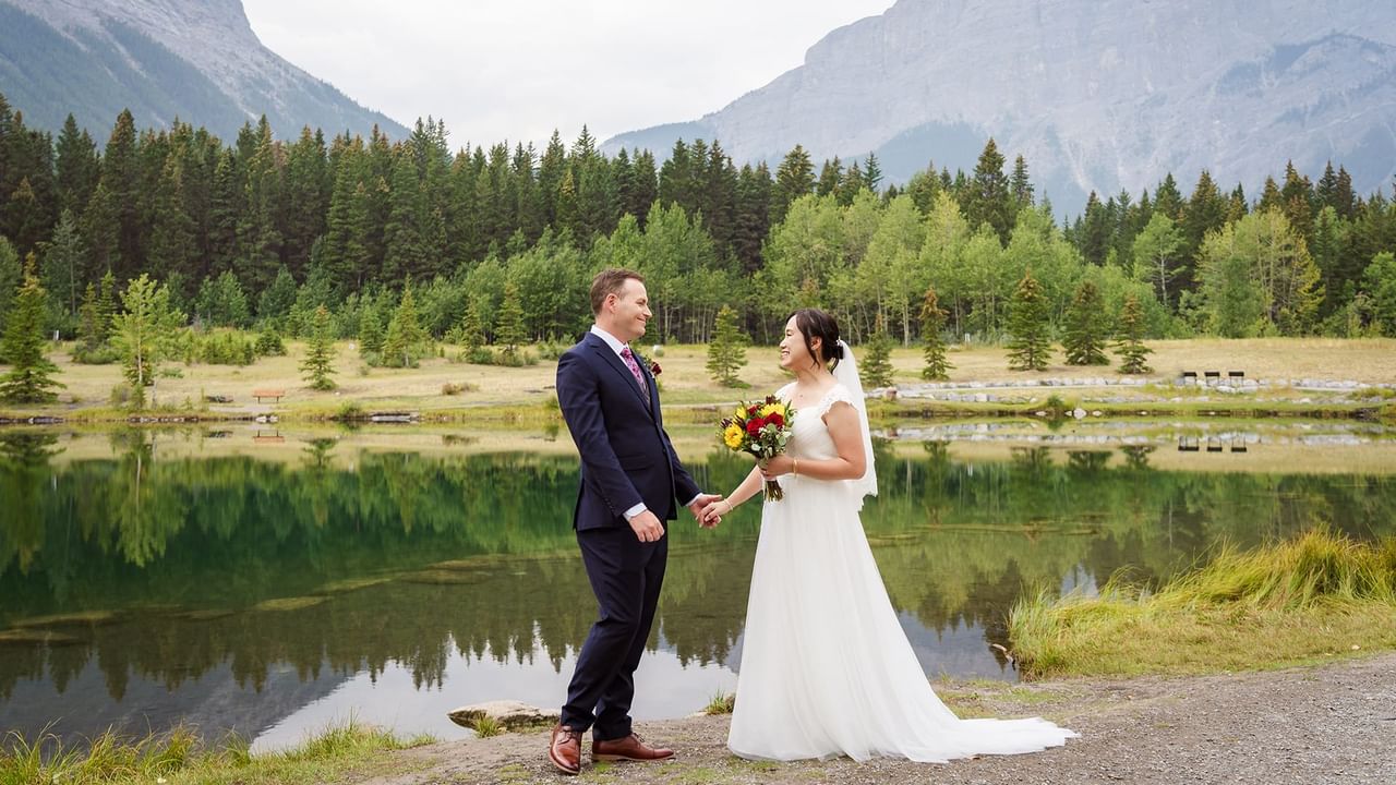 Newlyweds standing by a lake with mountain backdrop, holding hands, and smiling.