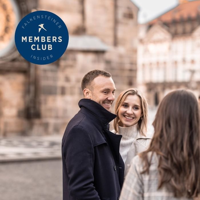 Troje nasmiješenih gostiju u razgovoru ispred povijesnih zgrada s logotipom Falkensteiner Members Club Insider