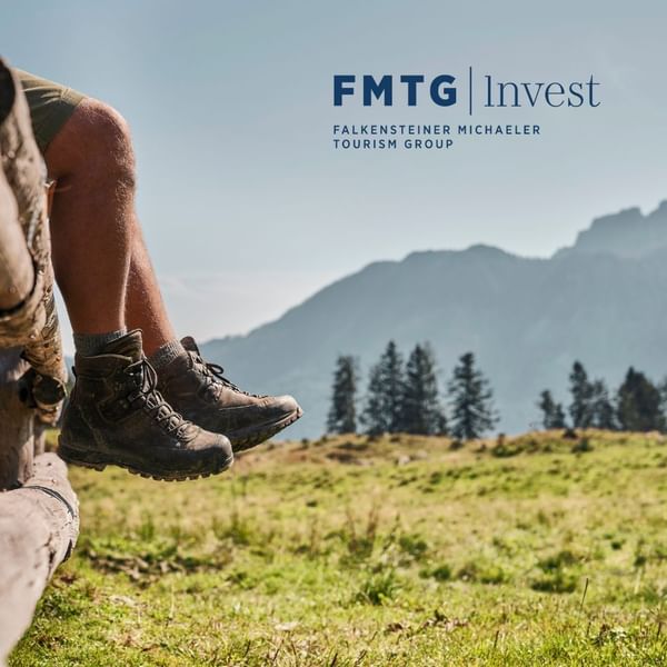 Persone sedute su un tronco in un prato con montagne sullo sfondo e logo FMTG Invest.