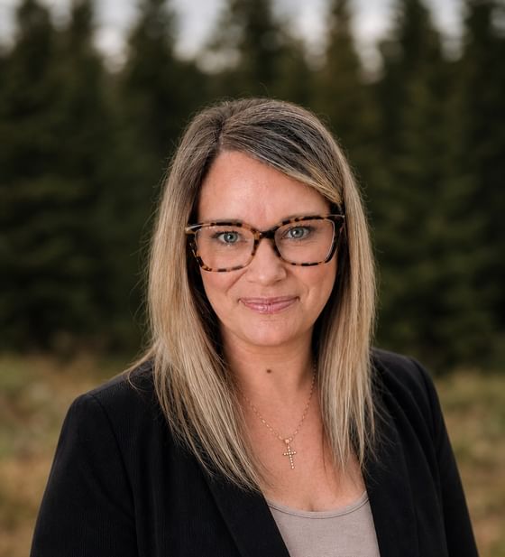 Lindi McKeown, wearing glasses and a black blazer, standing by a line of green trees at Oregon Garden Resort