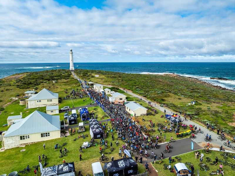 Aerial view of Cape to Cape MTB 2026 cyclists gathering near a coastal lighthouse with ocean views and green landscape.