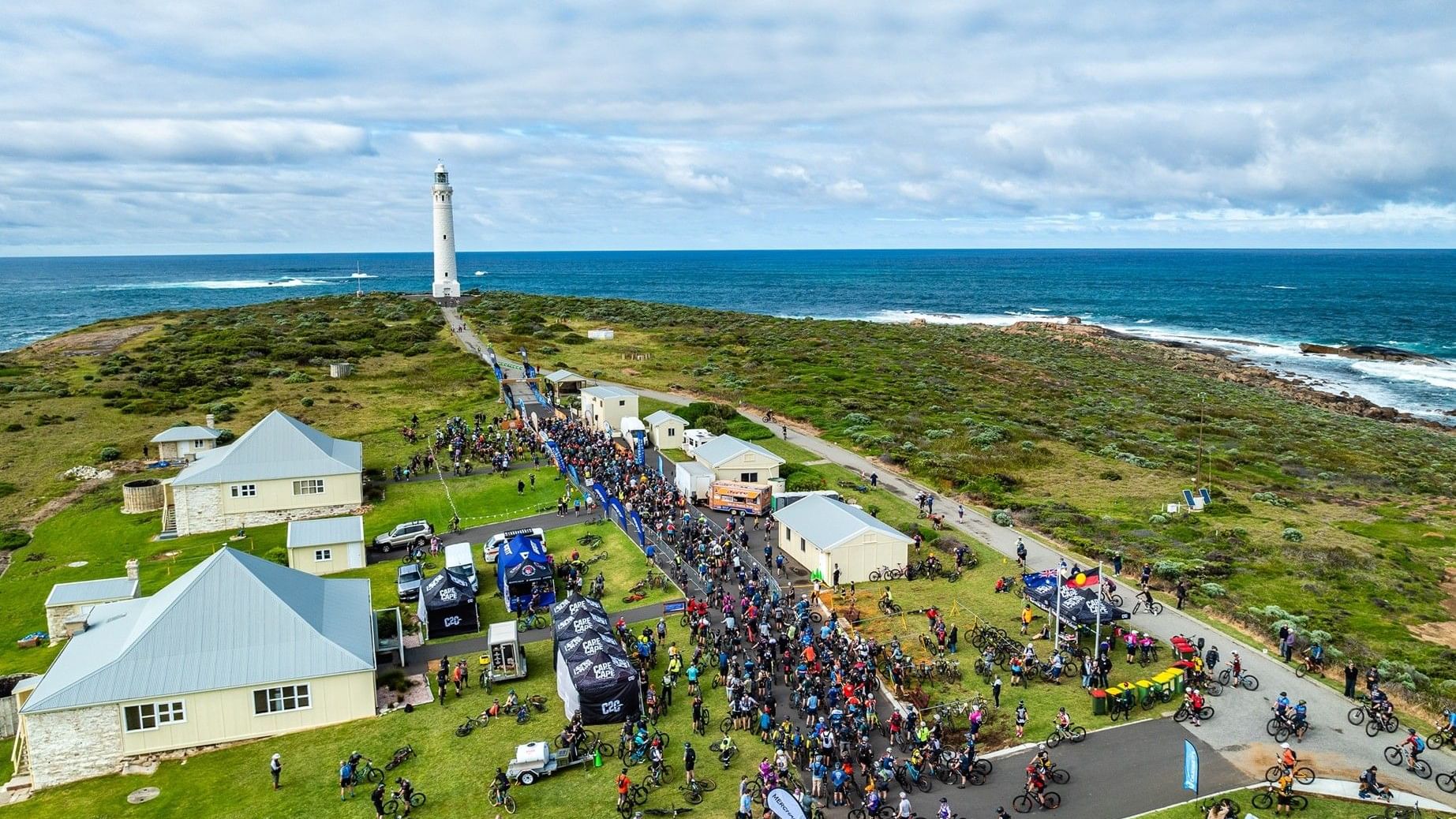 Aerial view of Cape to Cape MTB 2026 cyclists gathering near a coastal lighthouse with ocean views and green landscape.