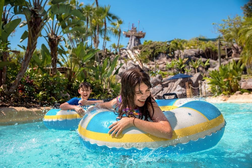 A boy and a girl in blue, yellow, and white inner tubes float in a lazy river at Typhoon Lagoon with tropical landscaping behind them and a boat stuck on a mountain peak in the distance.