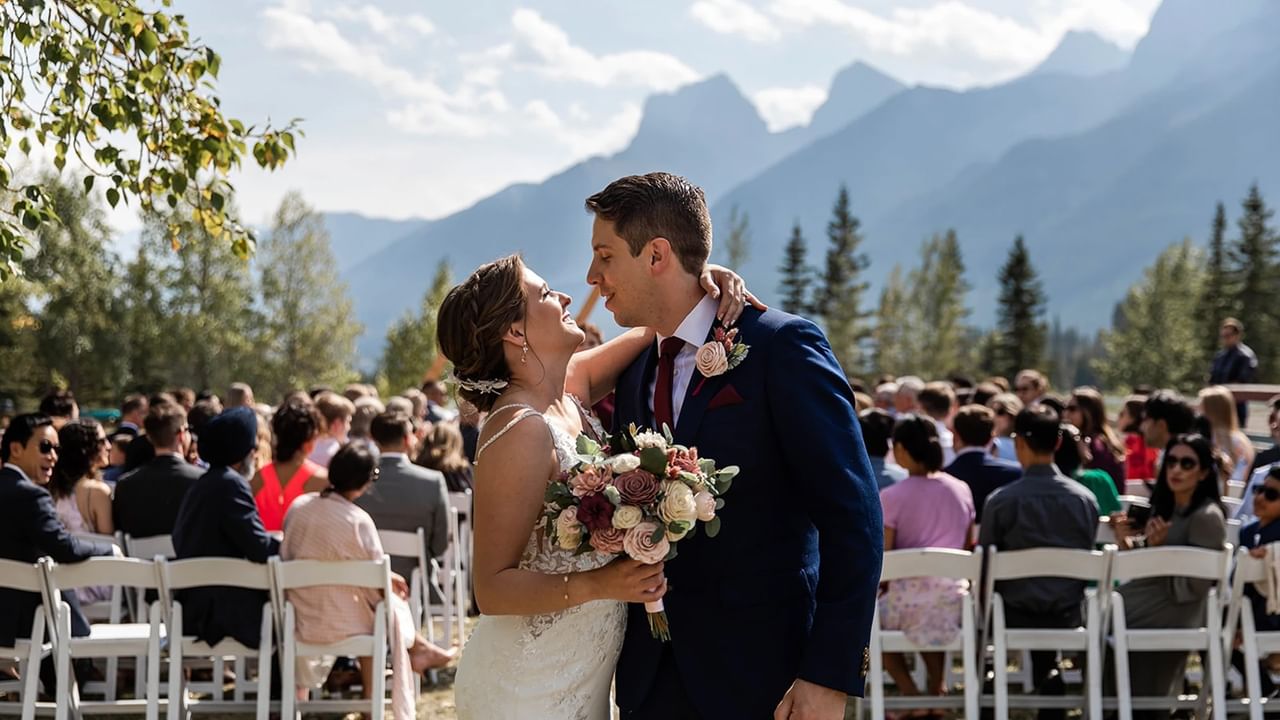 Bride and groom share a kiss at an outdoor wedding ceremony with mountains in the background.